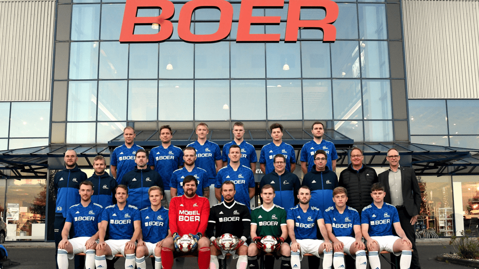 Mannschaftsbild einer Fußballmannschaft in blauen Trikots vor einem Geschäft mit "BOER"-Schild im Hintergrund.