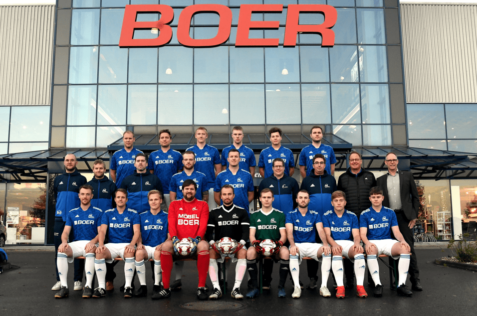 Mannschaftsbild einer Fußballmannschaft in blauen Trikots vor einem Geschäft mit "BOER"-Schild im Hintergrund.