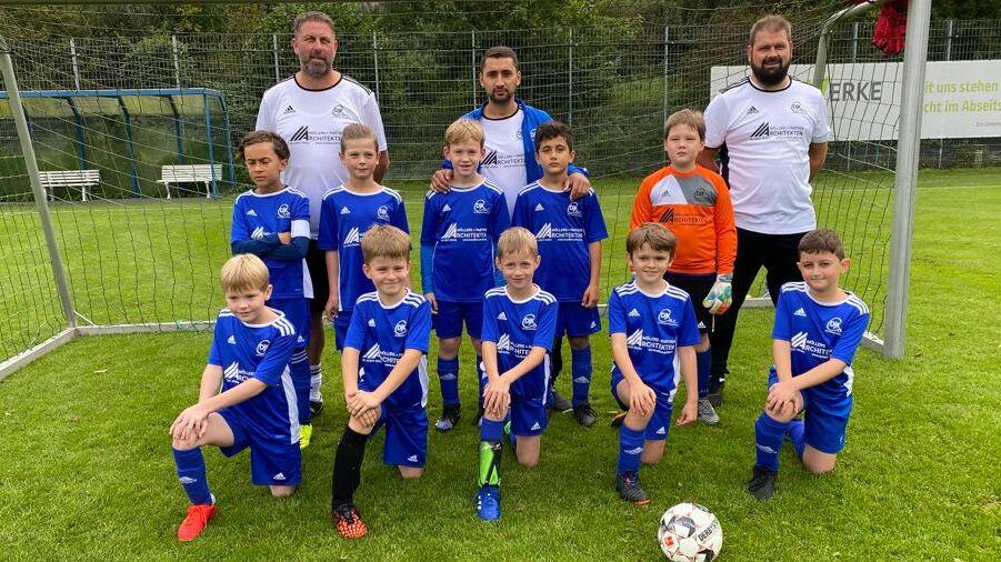 Gruppe von Kindern in blauen Fu&szlig;balltrikots posiert mit zwei Trainern vor einem Fu&szlig;balltor auf einem Rasenplatz.