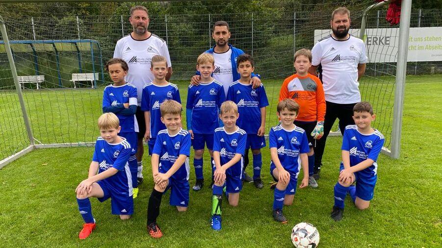 Gruppe von Kindern in blauen Fußballtrikots posiert mit zwei Trainern vor einem Fußballtor auf einem Rasenplatz.