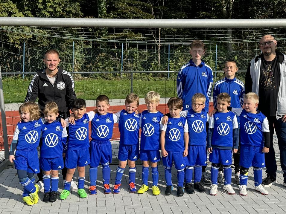 Gruppe von Kindern in blauen Fußballtrikots steht vor einem Tor, umgeben von zwei Betreuern auf einem Sportplatz.