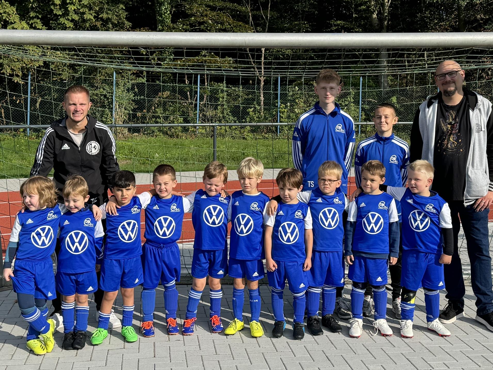 Gruppe von Kindern in blauen Fußballtrikots steht vor einem Tor, umgeben von zwei Betreuern auf einem Sportplatz.