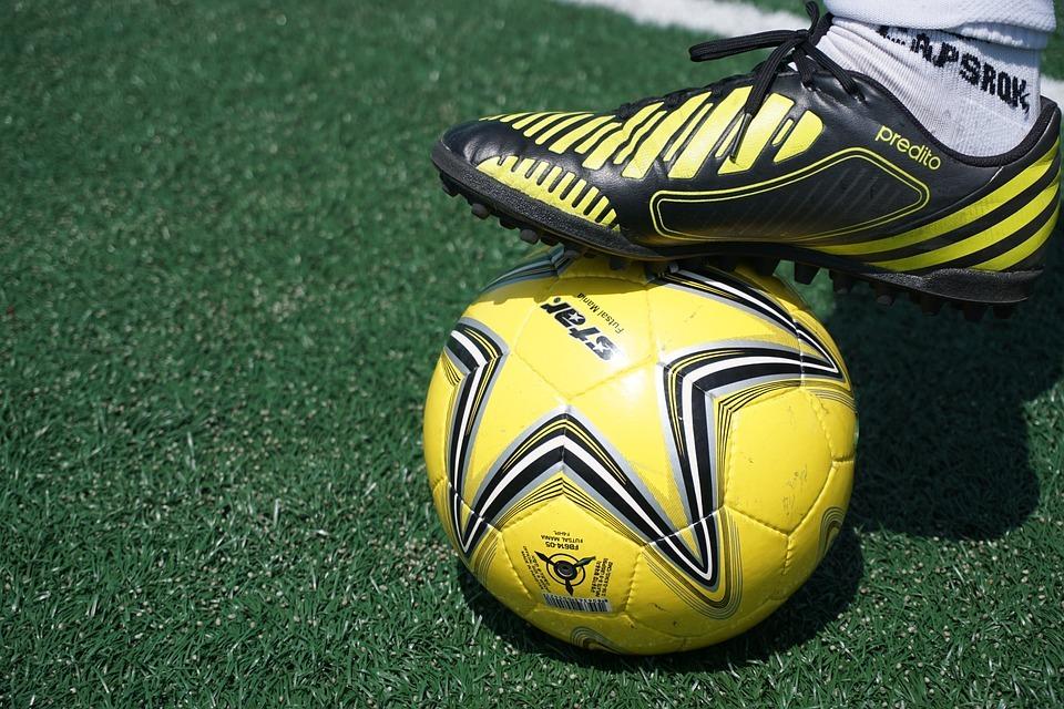 Schwarzer Fußballschuh mit gelben Streifen auf einem gelben Fußball, der auf grünem Kunstrasen liegt.