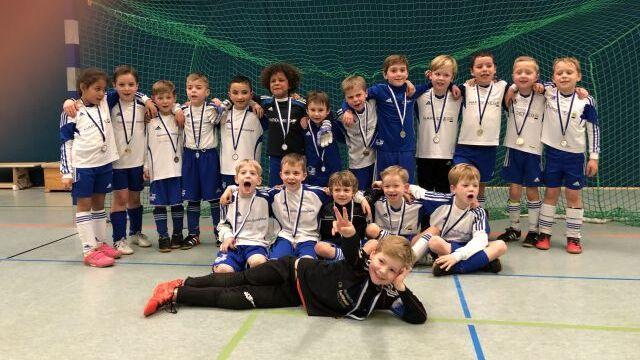 Gruppe von 20 Kindern in weißen und blauen Sportanzügen mit Medaillen, lächelnd in einem Sporthallen-Set.