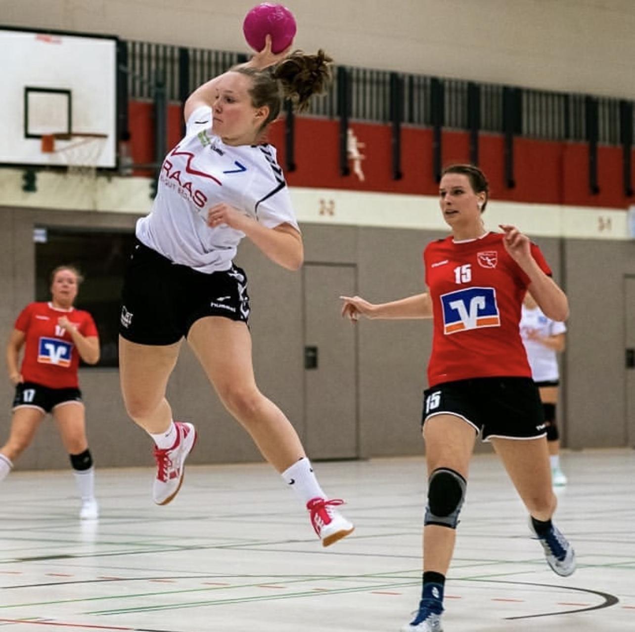 Sprungende Handballspielerin mit pinkem Ball, während eine Gegenspielerin in roter Uniform näherkommt.