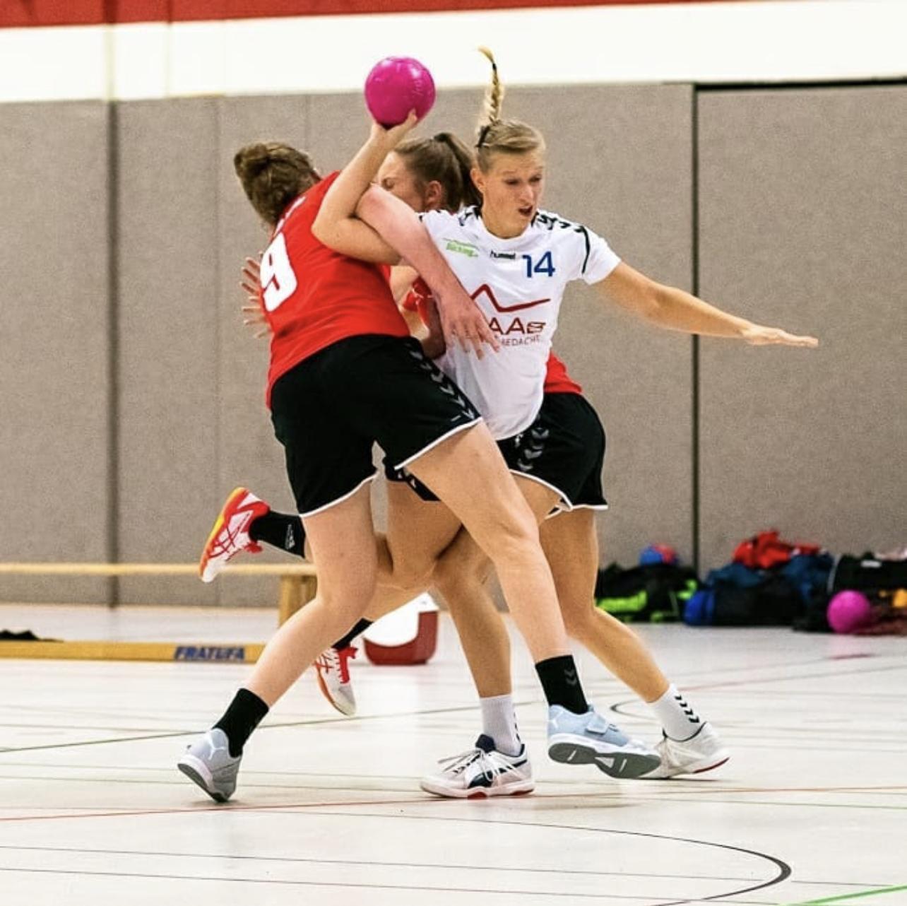 Drei Handballspielerinnen in Aktion, während eine von ihnen einen pinken Ball wirft und von zwei anderen Spielern gestoppt wird.