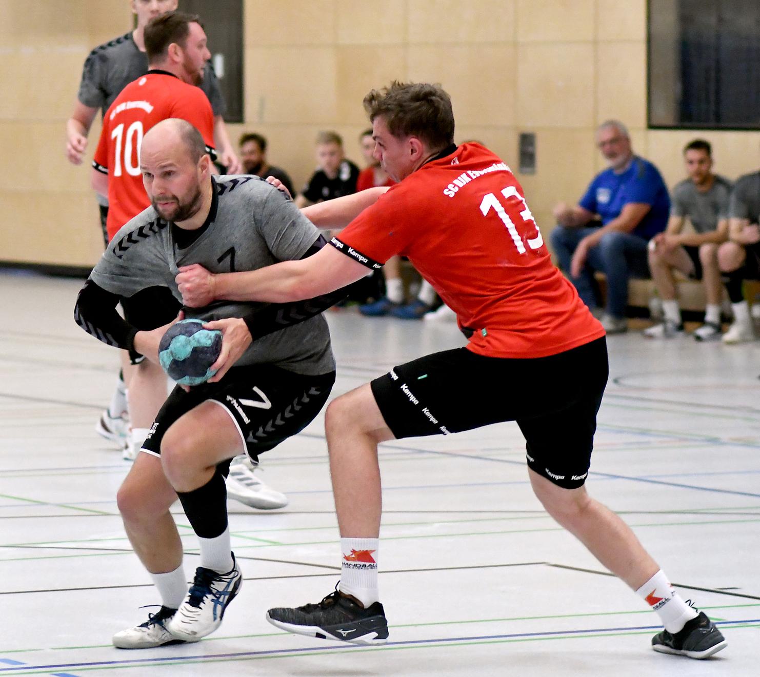 Zwei Handballspieler in Aktion; einer mit Ball, der andere greift ihn an, während Zuschauer im Hintergrund sitzen.
