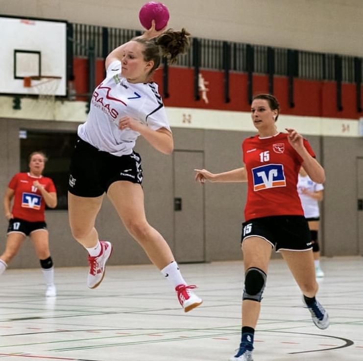 Sprungende Handballspielerin mit pinkem Ball, während eine Gegenspielerin in roter Uniform näherkommt.
