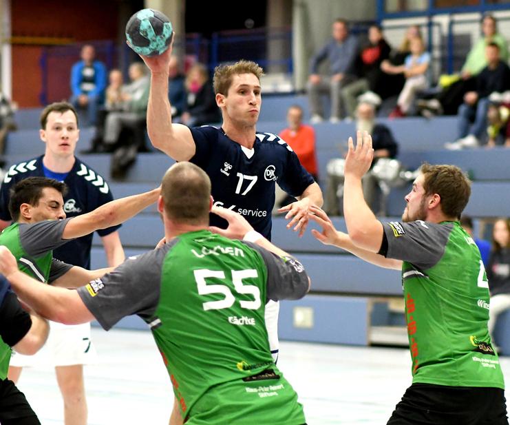 Spielszene im Handball: Spieler wirft den Ball, während zwei Verteidiger ihn abwehren möchten. Zuschauer im Hintergrund.