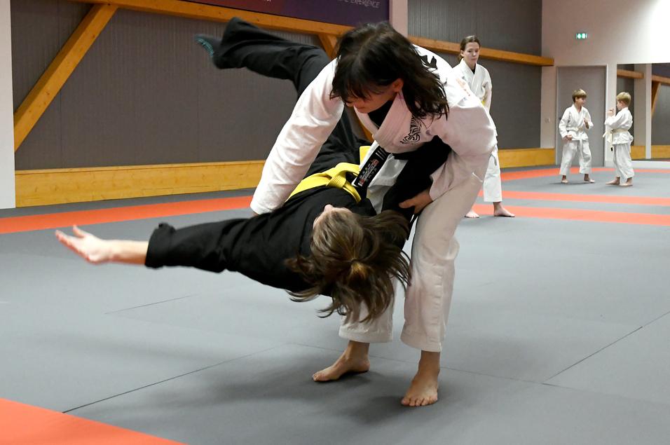Zwei Judokas üben einen Wurf auf einer Matte, während weitere Judokas im Hintergrund trainieren.