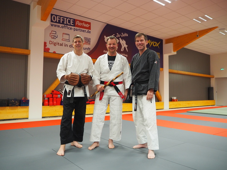 Drei Männer in Kampfbekleidung stehen auf einer Matte in einem Trainingsraum für Ju-Jutsu.