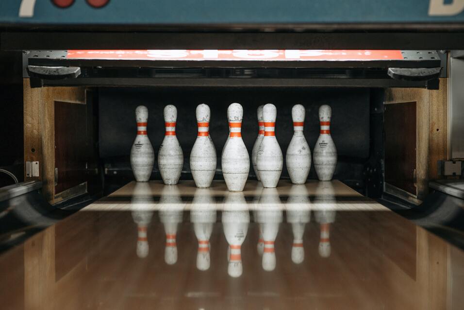 Sieben Bowlingpins mit orangefarbenen Streifen stehen auf einer Bowlingbahn. Reflektionen sind deutlich sichtbar.