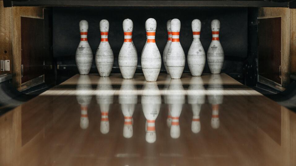 Sieben Bowlingpins mit orangefarbenen Streifen stehen auf einer Bowlingbahn. Reflektionen sind deutlich sichtbar.