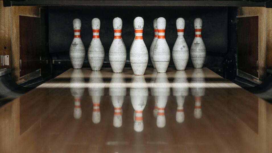 Sieben Bowlingpins mit orangefarbenen Streifen stehen auf einer Bowlingbahn. Reflektionen sind deutlich sichtbar.