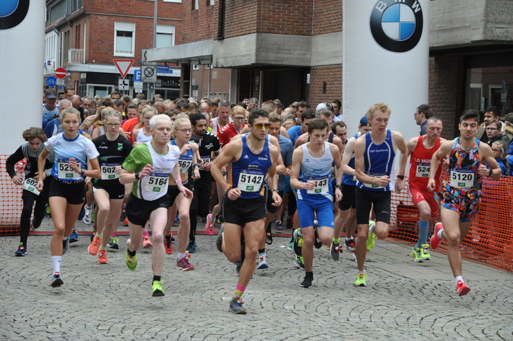 Start eines Laufwettbewerbs mit zahlreichen Teilnehmern in Sportkleidung, auf einer gepflasterten Straße.