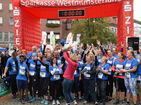 Gruppierung von Läufern in blauen Shirts feiert am Startbogen eines Stadtlaufs mit ausgestreckten Händen und jubelndem Publikum.
