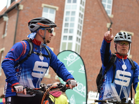 Zwei Radfahrer in blauen Trikots stehen an einem Veranstaltungsort, einer zeigt mit dem Finger in eine Richtung.
