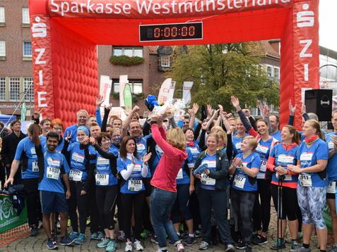 Gruppierung von L&auml;ufern in blauen Shirts feiert am Startbogen eines Stadtlaufs mit ausgestreckten H&auml;nden und jubelndem Publikum.