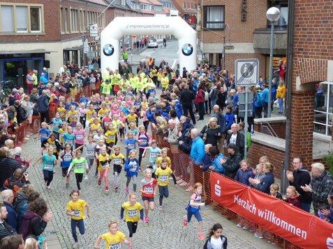 Kinderlauf mit Teilnehmern in bunten T-Shirts, umgeben von Zuschauern und einer festlichen Atmosphäre auf der Straße.