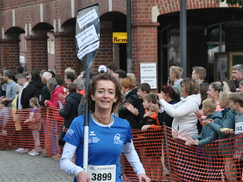 Läuferin in blauer Sportbekleidung trägt ein Schild und läuft auf einem Wettkampf zu einer zuschauenden Menge.