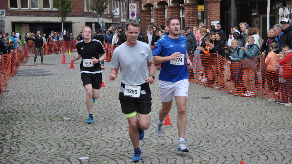 Drei Läufer in Sportkleidung absolvieren ein Rennen, von Zuschauern und Absperrungen umgeben.
