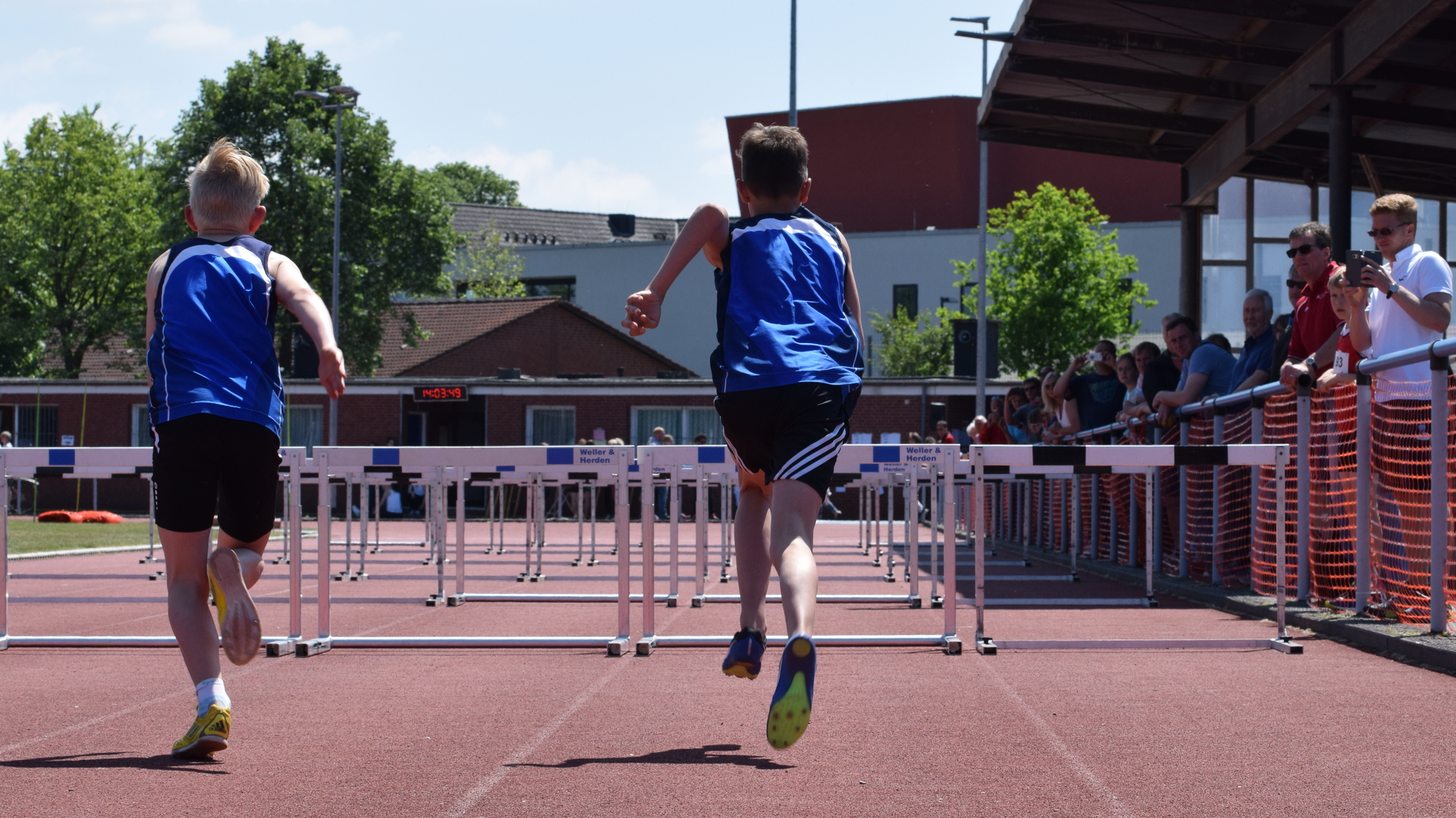 Zwei Jungen in blauen Trikots sprinten &uuml;ber H&uuml;rden auf einer roten Laufbahn, Zuschauer an der Seite beobachten das Rennen.