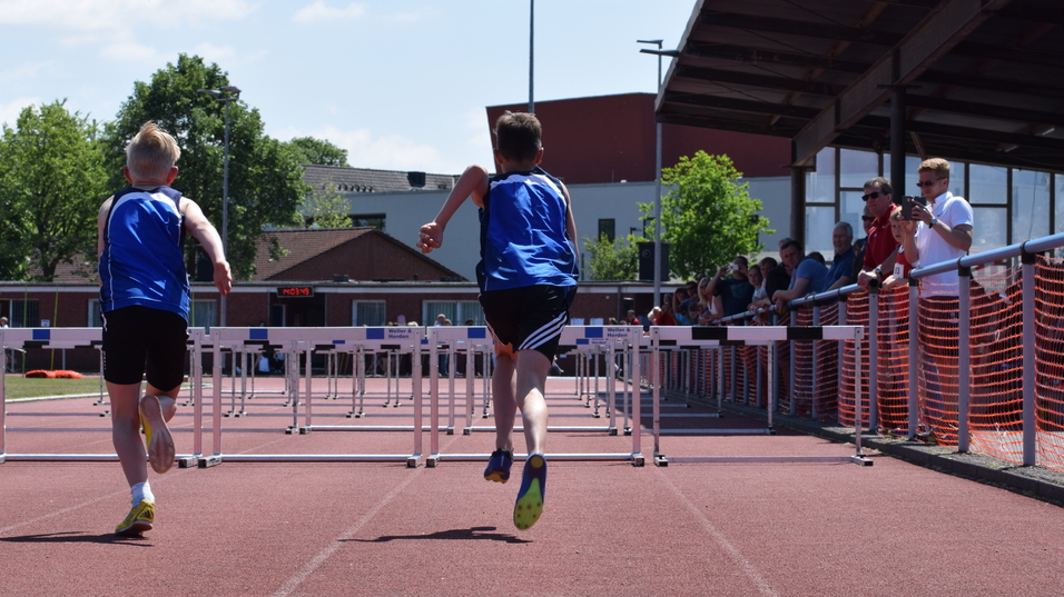 Zwei Jungen in blauen Trikots sprinten über Hürden auf einer roten Laufbahn, Zuschauer an der Seite beobachten das Rennen.
