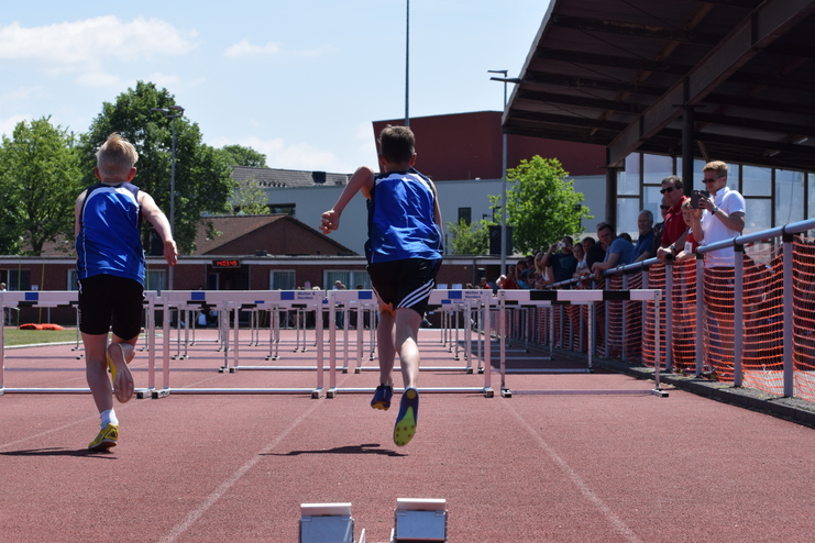 Zwei Jungen in blauen Trikots sprinten über Hürden auf einer roten Laufbahn, Zuschauer an der Seite beobachten das Rennen.