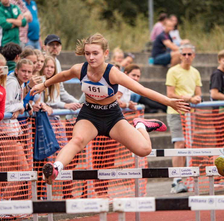 Eine Athletin springt über eine Hürde bei einem Leichtathletik-Wettbewerb, umgeben von Zuschauerinnen und Zuschauern.