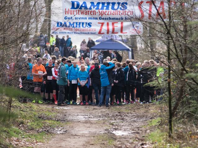 Starterfeld mit Teilnehmern vor dem Banner "START" im Wald bei einem Laufereignis. B&auml;ume und ein Weg im Vordergrund.