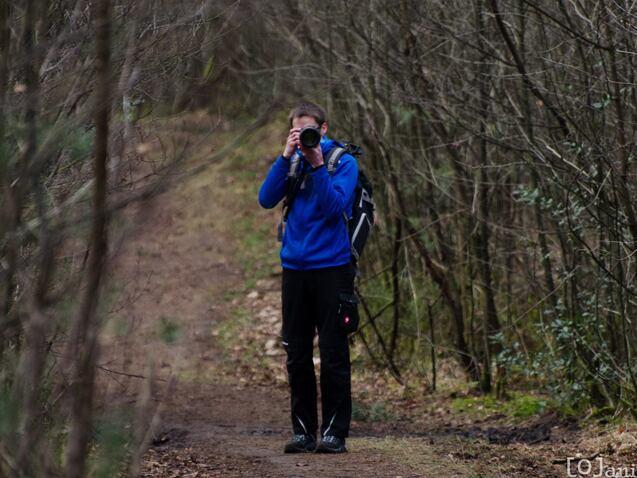 Ein Fotograf in blauer Jacke hält eine Kamera auf einem Waldweg zwischen Bäumen und Sträuchern.
