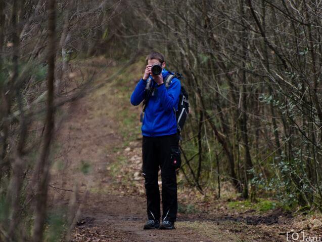 Ein Fotograf in blauer Jacke h&auml;lt eine Kamera auf einem Waldweg zwischen B&auml;umen und Str&auml;uchern.