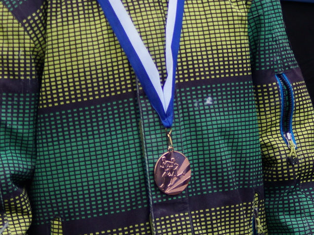 Junge mit Medaille in grüner Jacke steht vor einer Veranstaltung, neben ihm ein weiterer Teilnehmer in blauer Jacke.