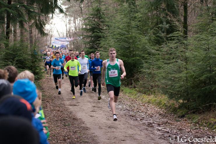 Läufer in Sportkleidung im Wettkampf auf einem geschotterten Waldweg zwischen Bäumen, Zuschauer am Rand der Strecke.