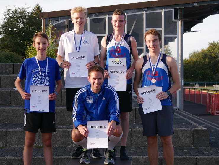 Gruppe von fünf Athleten mit Urkunden und Medaillen auf einer Treppe, hinter ihnen ein Sportplatz mit Tribüne.