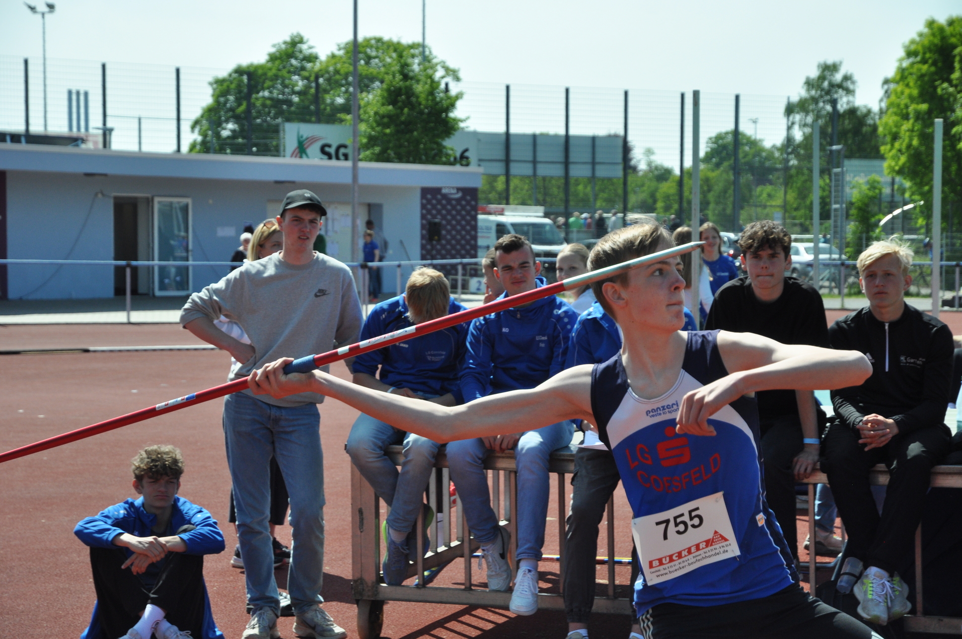 Junger Athlet mit Speer in der Werferpose, umgeben von zuschauenden Sportlern auf einem roten Sportplatz.