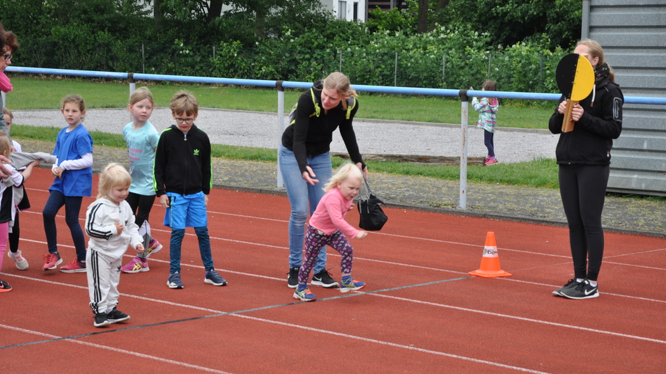 Kinder laufen auf einer Leichtathletikbahn, eine Betreuerin hält ein Schild, während eine Mutter ein Kind unterstützt.