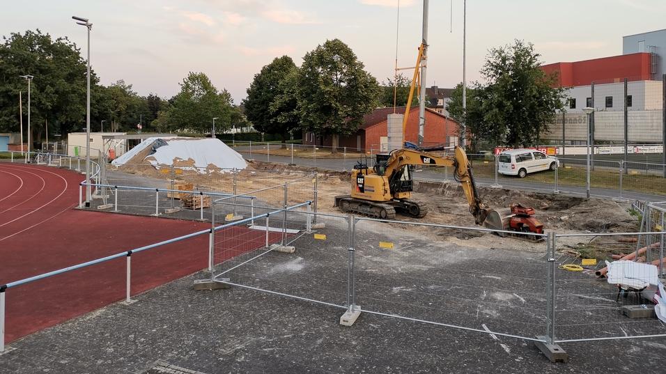 Baustelle mit Bagger und Kran, umgeben von Bauzaun, im Hintergrund eine leicht hügelige Landschaft und Sporteinrichtungen.