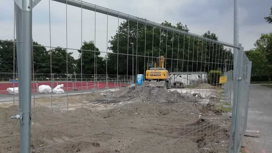 Bauzaun um ein Baustellengel&auml;nde mit Erdarbeiten und einem Bagger im Hintergrund, unter bew&ouml;lktem Himmel.