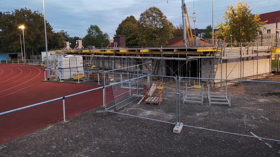 Bauarbeiten an einem Sportgebäude mit Gerüsten und Baustellenmaterial im Hintergrund eines Sportplatzes.