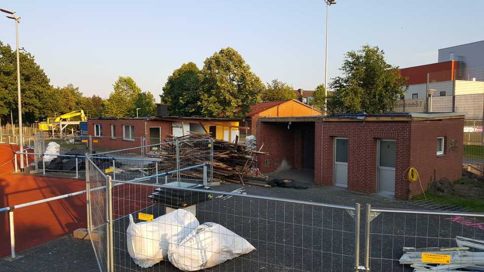 Bauarbeiten an mehreren roten Backsteingebäuden mit Holzstapel und Bauzäunen auf einem Sportgelände.