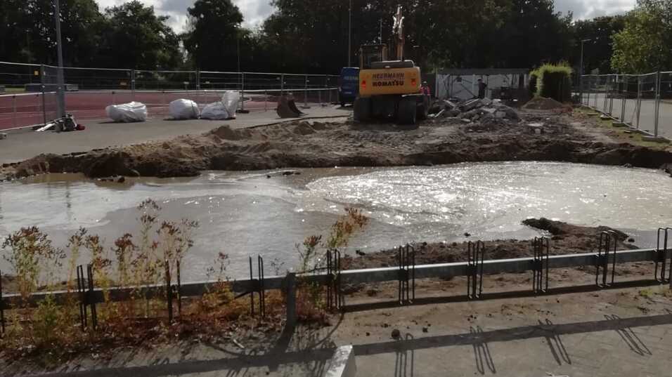 Baustelle mit Bagger auf einer Erdebene, umgeben von Baustellenzaun und Büschen unter bewölktem Himmel.