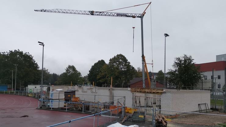 Bauarbeiten an einem Sportgel&auml;nde mit Kran, Baustellenmaterial und einem Mann in blauer T-Shirt im Vordergrund.
