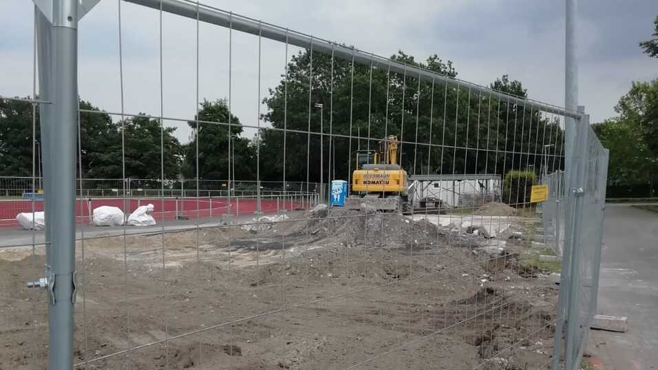 Bauzaun um ein Baustellengelände mit Erdarbeiten und einem Bagger im Hintergrund, unter bewölktem Himmel.