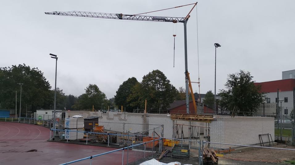Bauarbeiten an einem Sportgelände mit Kran, Baustellenmaterial und einem Mann in blauer T-Shirt im Vordergrund.