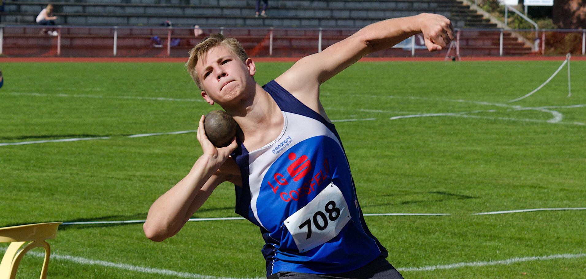 Jugendlicher Athlet im blauen Trikot wirft eine Kugel auf einem Sportplatz mit grüner Rasenfläche im Hintergrund.