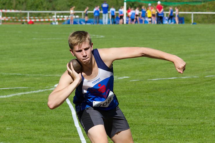 Junger Athlet beim Kugelstoßen auf einem grünen Sportfeld, konzentriert in der Wurfbewegung.