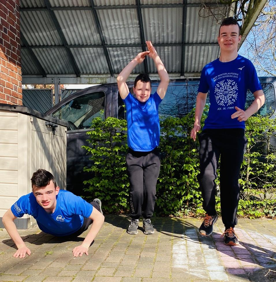 Drei Jungen in blauen T-Shirts machen sportliche Übungen draußen auf einem gepflasterten Boden.