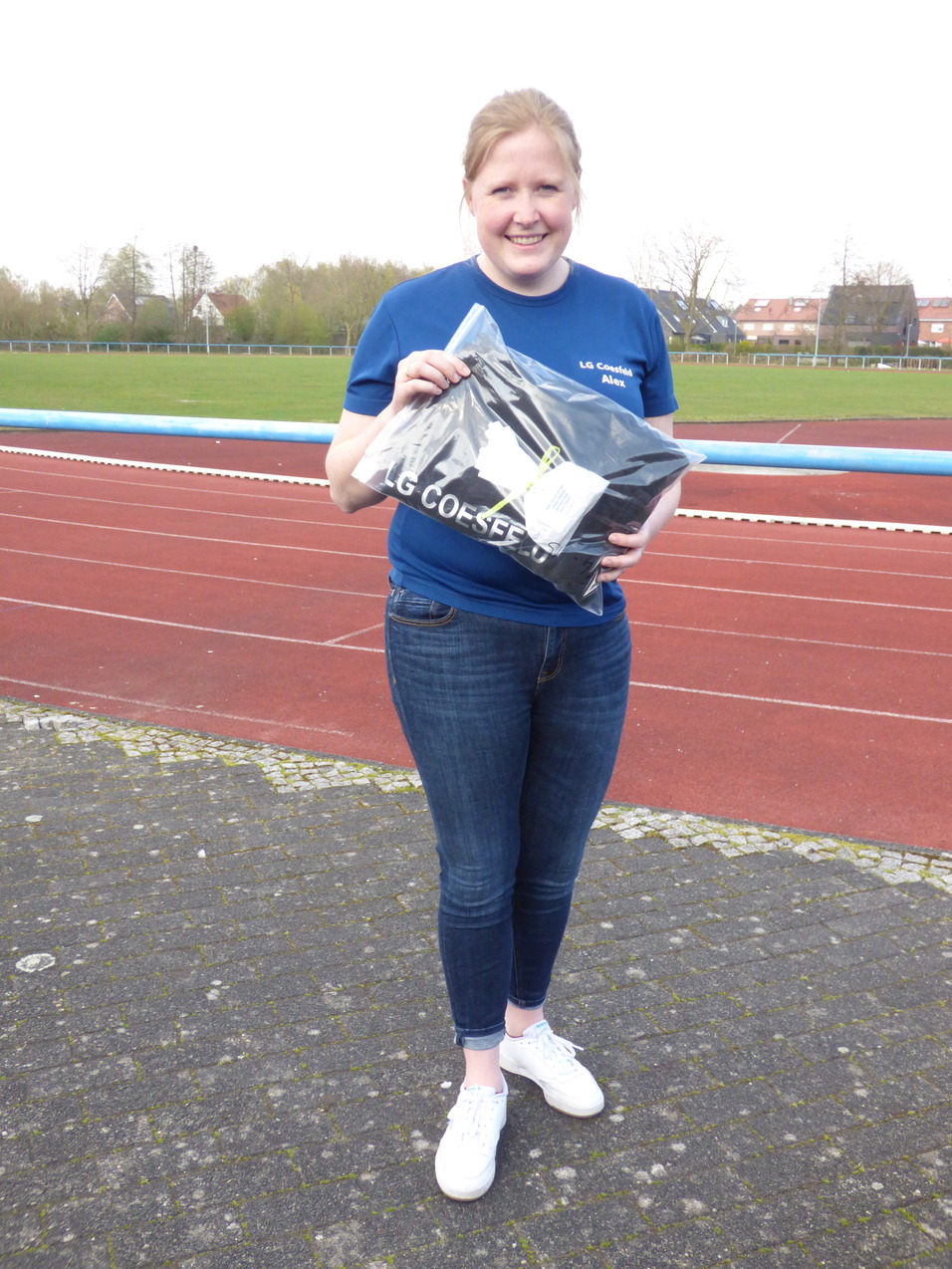 Frau auf einer Laufbahn hält ein T-Shirt in einer Plastiktüte, trägt ein blaues Shirt und Jeans, lächelt.
