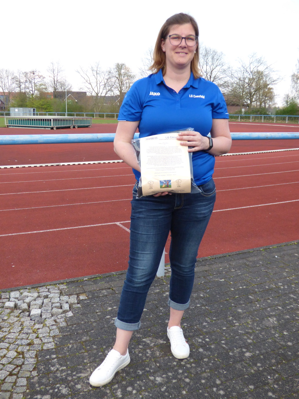 Frau mit Brille in blauem Sportshirt hält ein Dokument vor einer Laufbahn im Stadion in der Nähe von Bäumen.